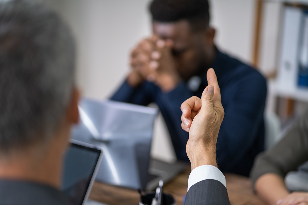A person pointing their finger aggressively at a colleague across a desk, while the colleague sits with their head down in a distressed posture, conveying workplace bullying or intimidation.
