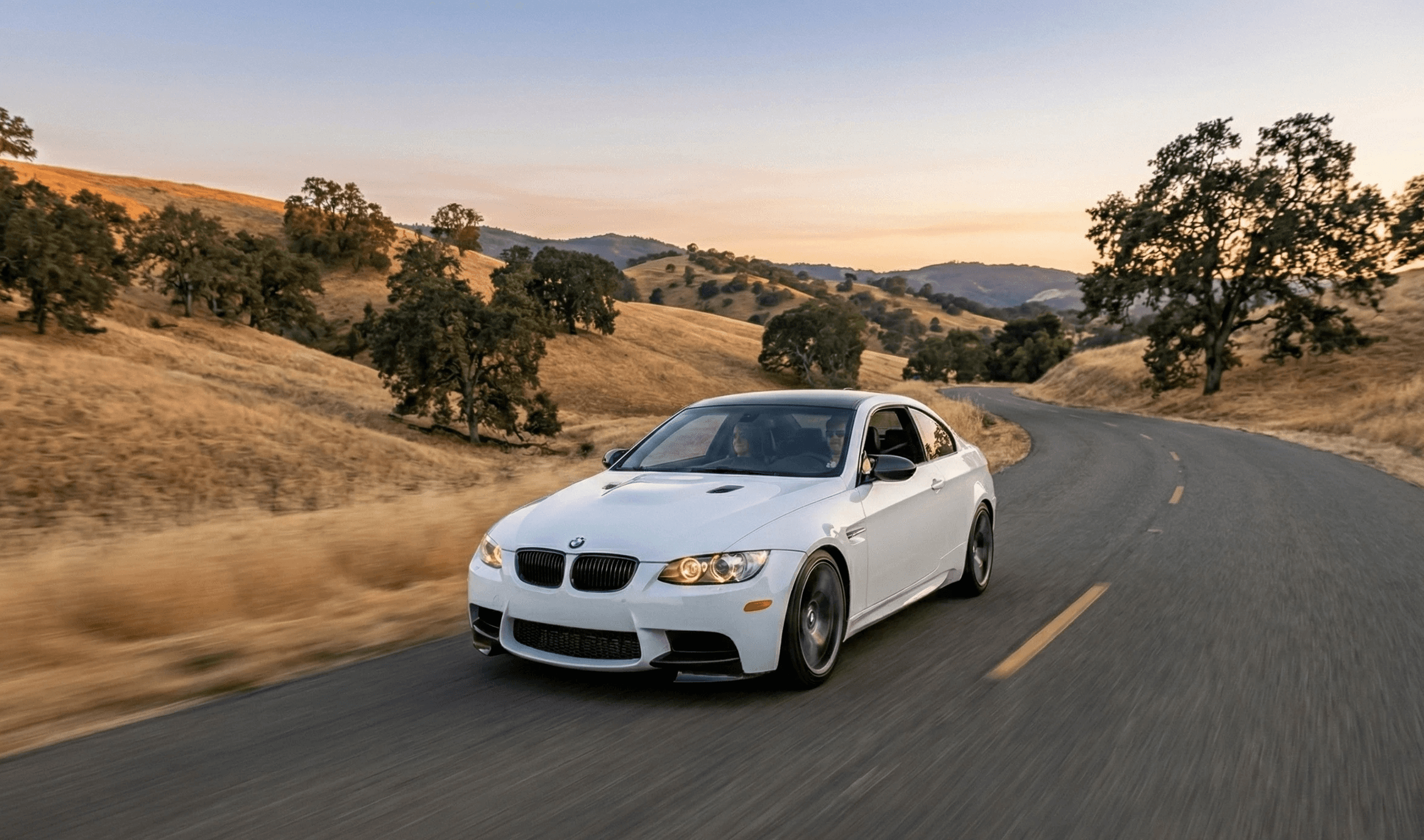 White BMW E92 M3 coupe driving on scenic winding road at golden hour, highlighting pristine condition for enthusiast car resale value