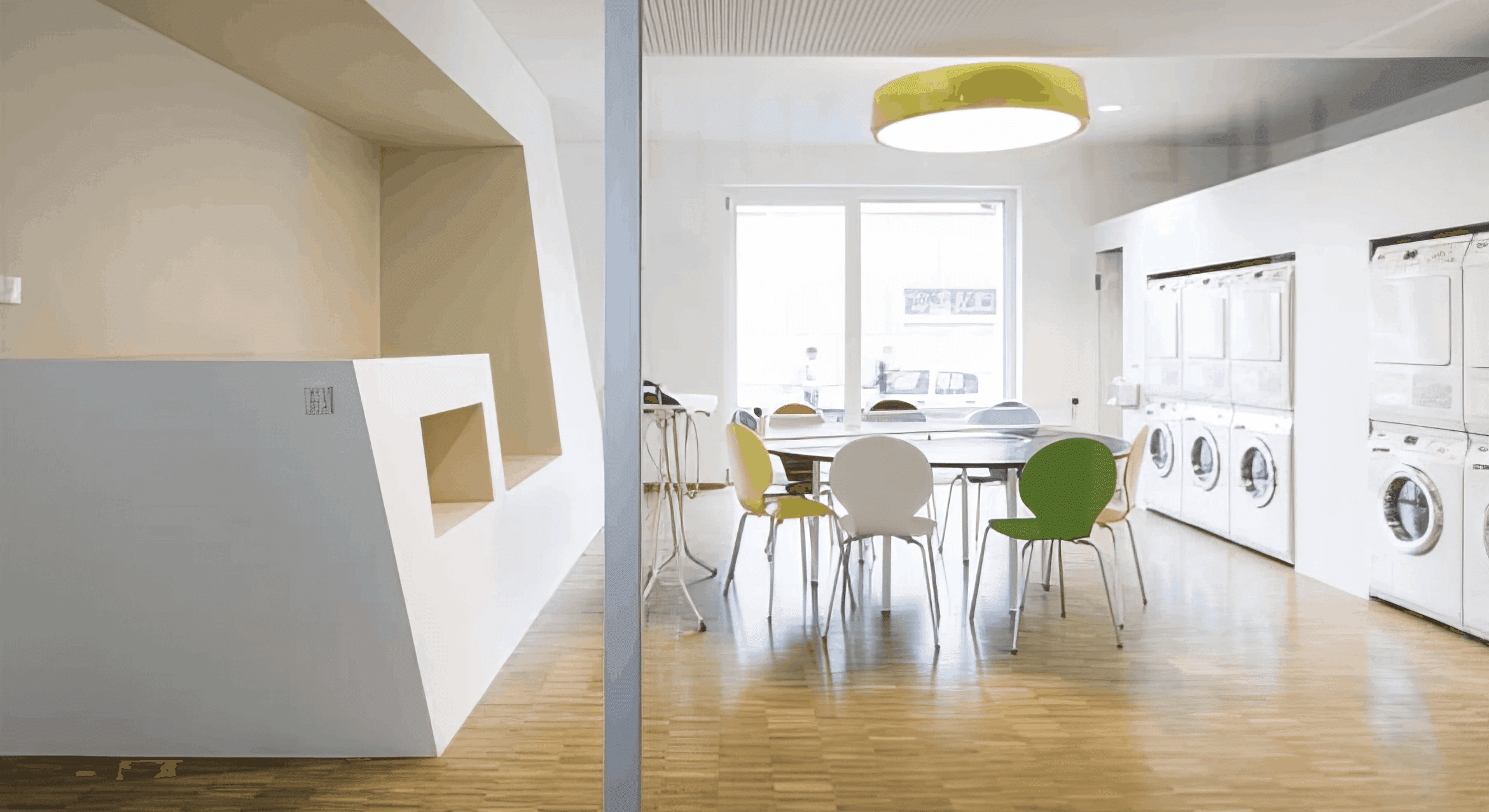 MW Interior - Heller Gemeinschaftsraum mit bunten Schalenstühlen in Gelb, Grün, Weiß und Orange, rundem Tisch, gelber Pendelleuchte und angrenzenden Waschmaschinen" Beschreibung: Farbenfroher Gemeinschaftsbereich mit bunten Stühlen und integriertem Waschsalon nebenan – ein Ort, der praktische Alltagsfunktion und soziale Begegnung spielend miteinander verbindet.