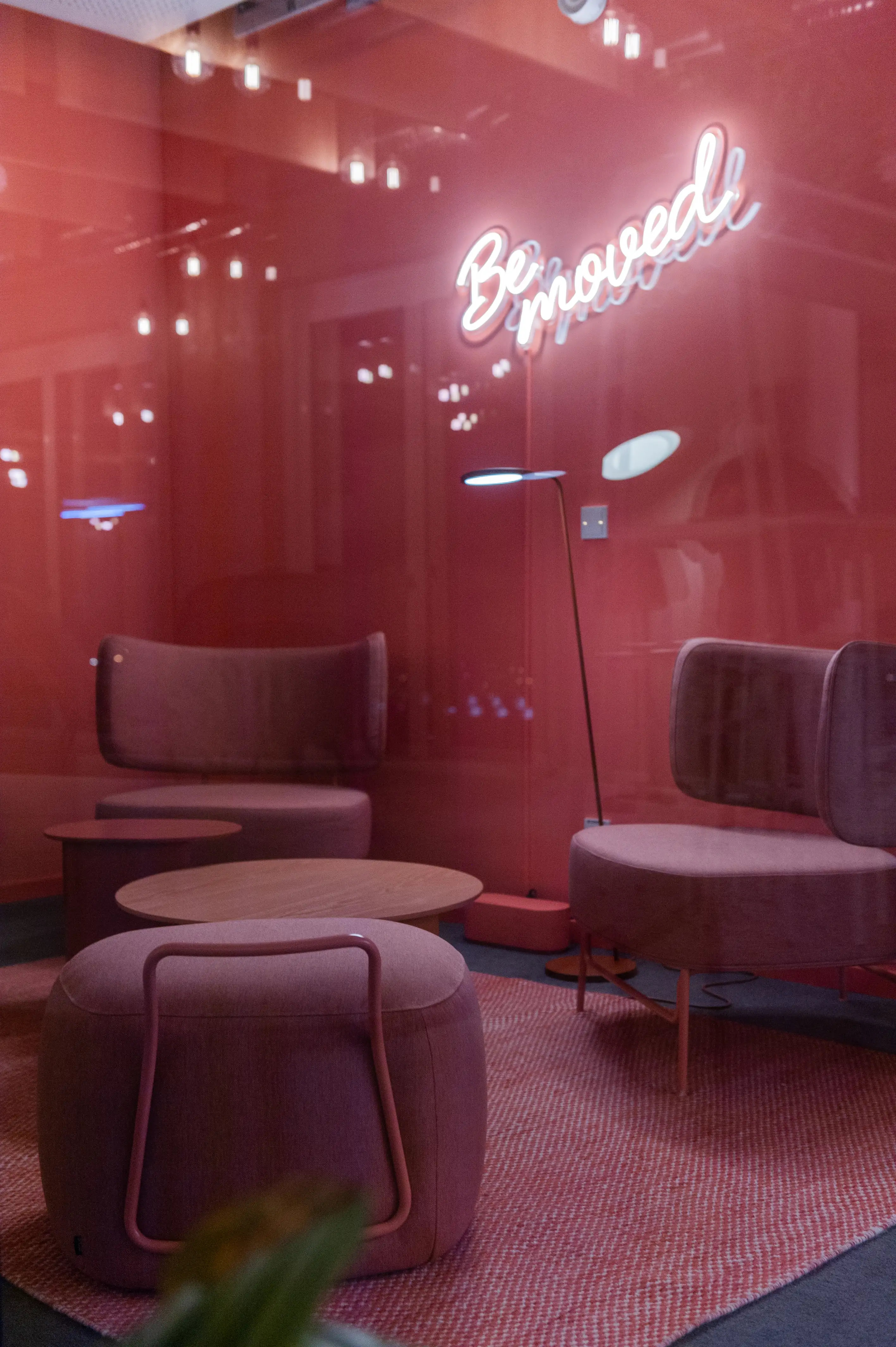 Coral-red lounge room at OMIO seen through glass, with a neon Be moved sign on the wall, dusty-pink upholstered chairs, round wooden coffee tables, a pouf and a woven red rug.