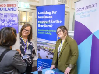 Three women engage in discussion at a business expo. Two stand by a booth with banners reading "Looking for business support in the Borders?" and "Success starts here."