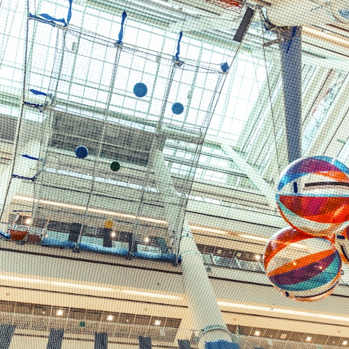 Colorful, large beach balls hang from the ceiling of a modern multi-story atrium with netting and glass skylights above.