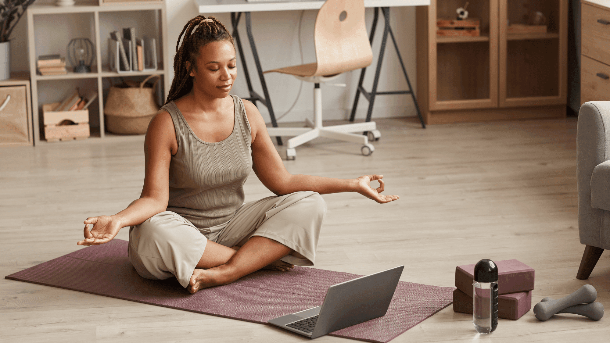 Woman practicing meditation in Easy Pose (Sukhasana) on a mauve yoga mat, following an online yoga class on laptop, with fitness equipment and yoga blocks nearby, demonstrating a hybrid approach to wellness