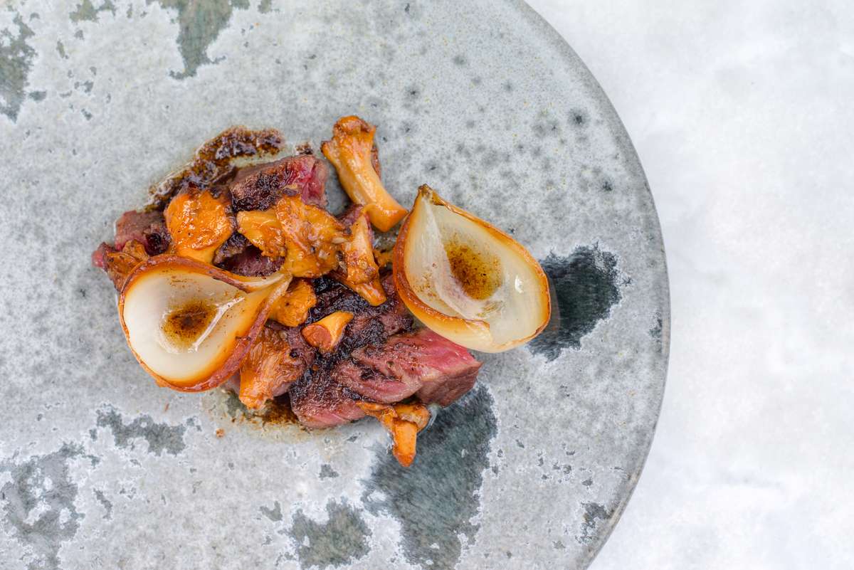 Seared meat and roasted vegetables with charred onion halves served on a textured gray ceramic plate.