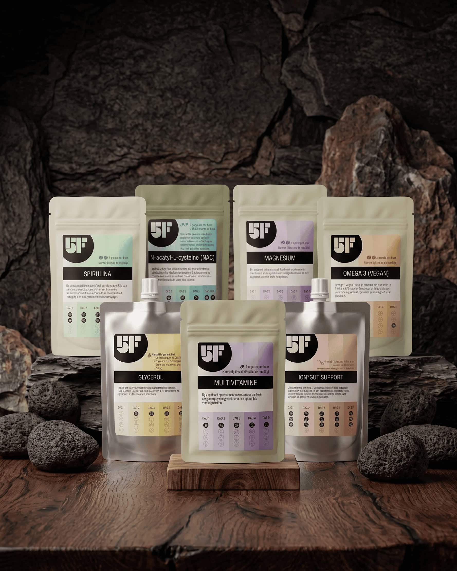 Zeven 5 Day Fast supplementenzakjes — Spirulina, NAC, Magnesium, Omega 3 Vegan, Glycerol, Ion Gut Support en Multivitamine — uitgestald op een houten ondergrond met donkere rotsen op de achtergrond.