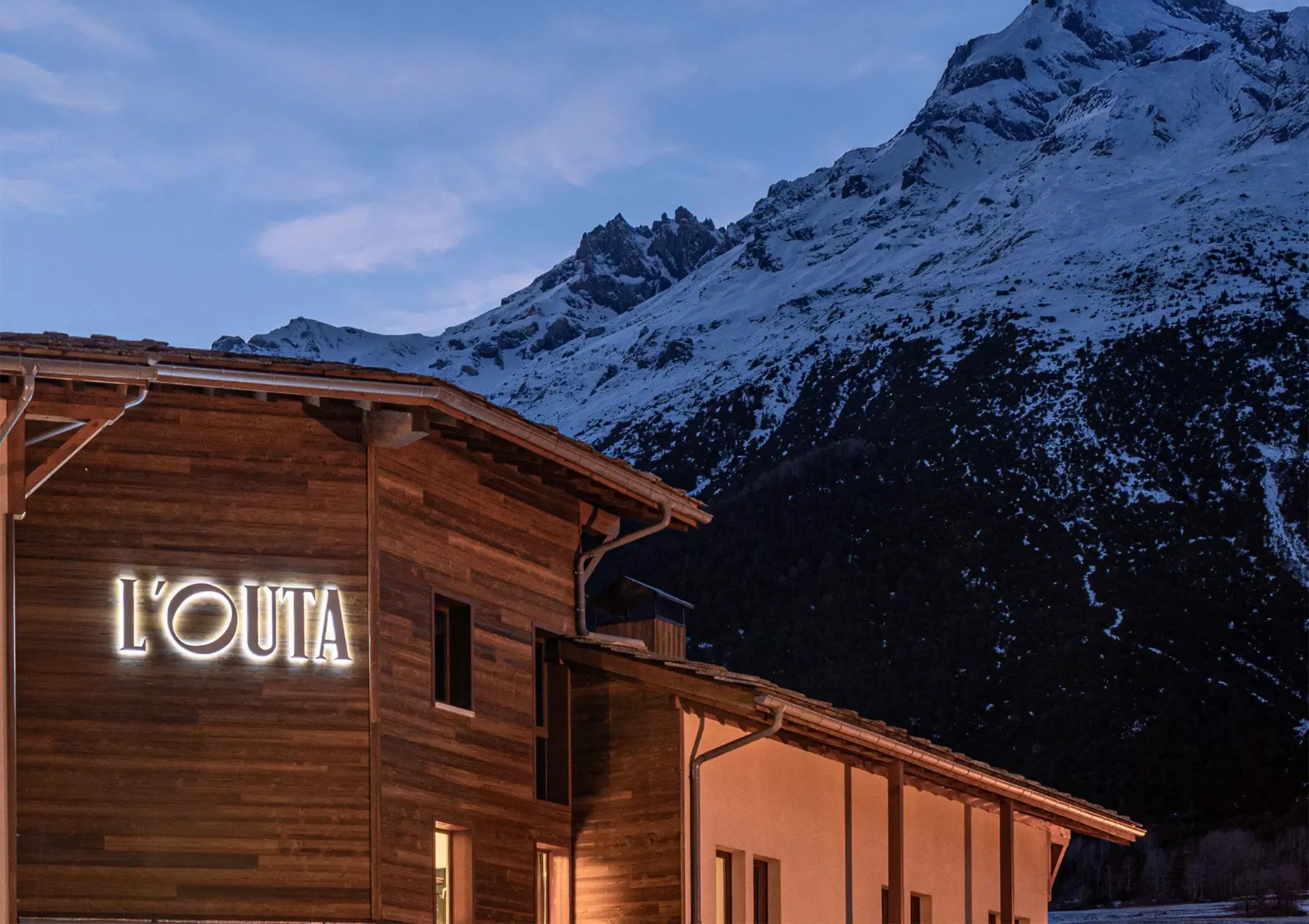 Mise en valeur de l'enseigne 'L'OUTA' sur un chalet moderne en bois sombre, contrastant avec le bleu du crépuscule et les montagnes de Savoie en arrière-plan.
