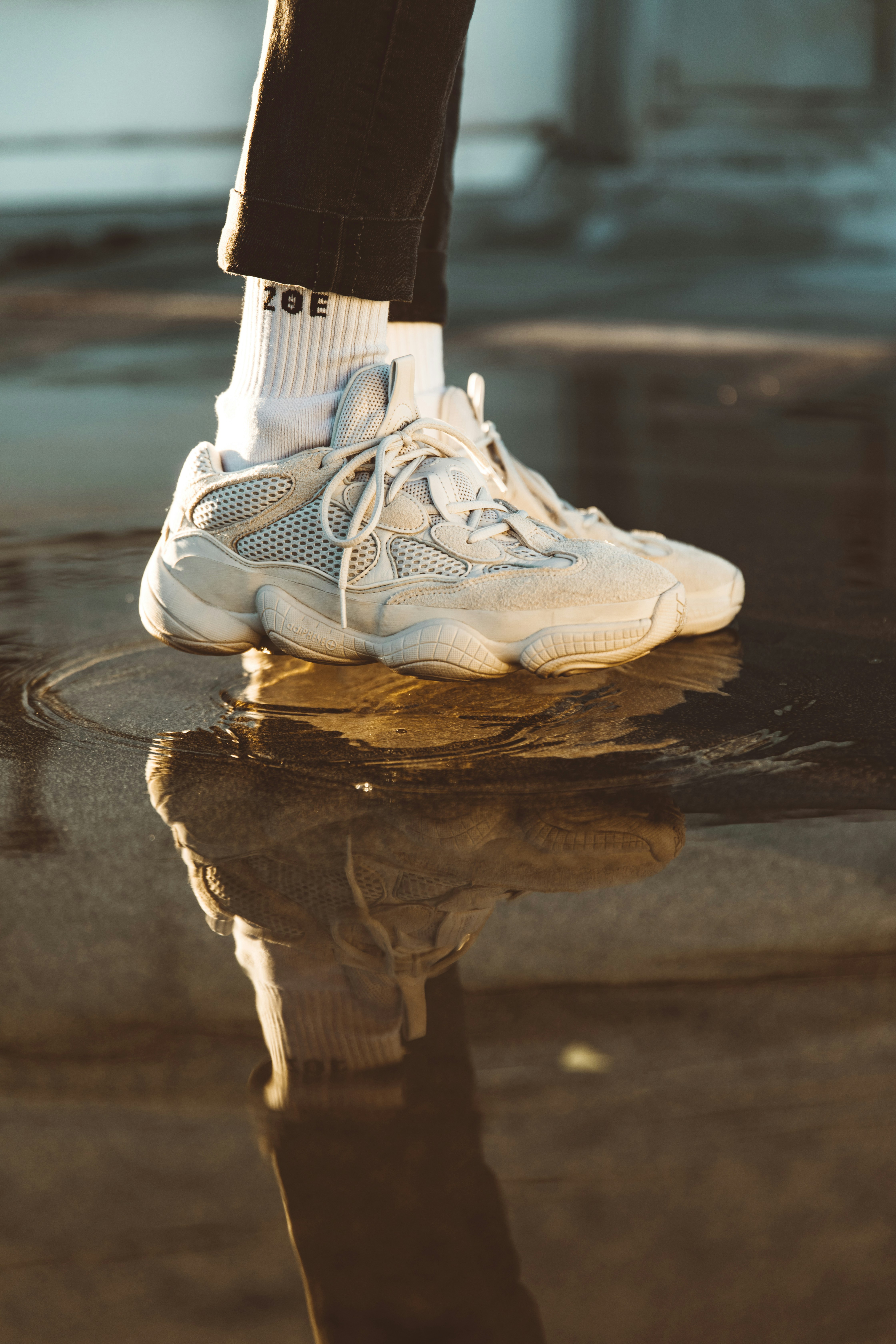 White sneaker stepping into a puddle on a city street, with the reflection of the wearer's leg visible in the water