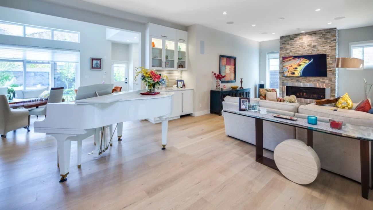 Open concept living and dining room in custom Vancouver home featuring white grand piano, light hardwood floors, and stone fireplace with modern design