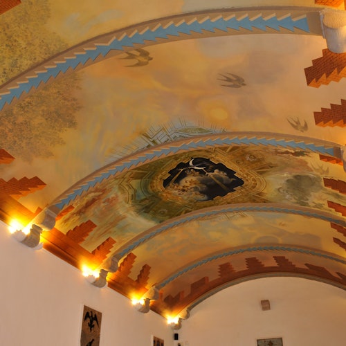 An ornate ceiling features colorful frescoes, including birds and architectural elements, illuminated by warm wall lights.