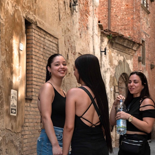 Three women stand chatting and smiling in a narrow street with weathered brick walls; one holds a bottle of water.