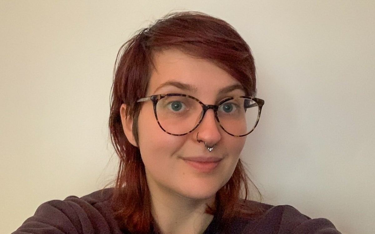 A person with short, reddish‑brown hair sits against a plain, light‑coloured background. They wear dark glasses and a casual top.