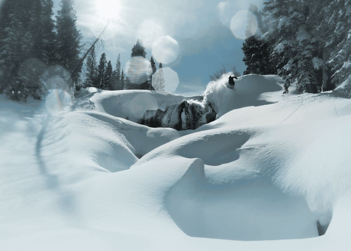 A skier launches off a snow-covered cliff above a frozen waterfall, framed by sun flare