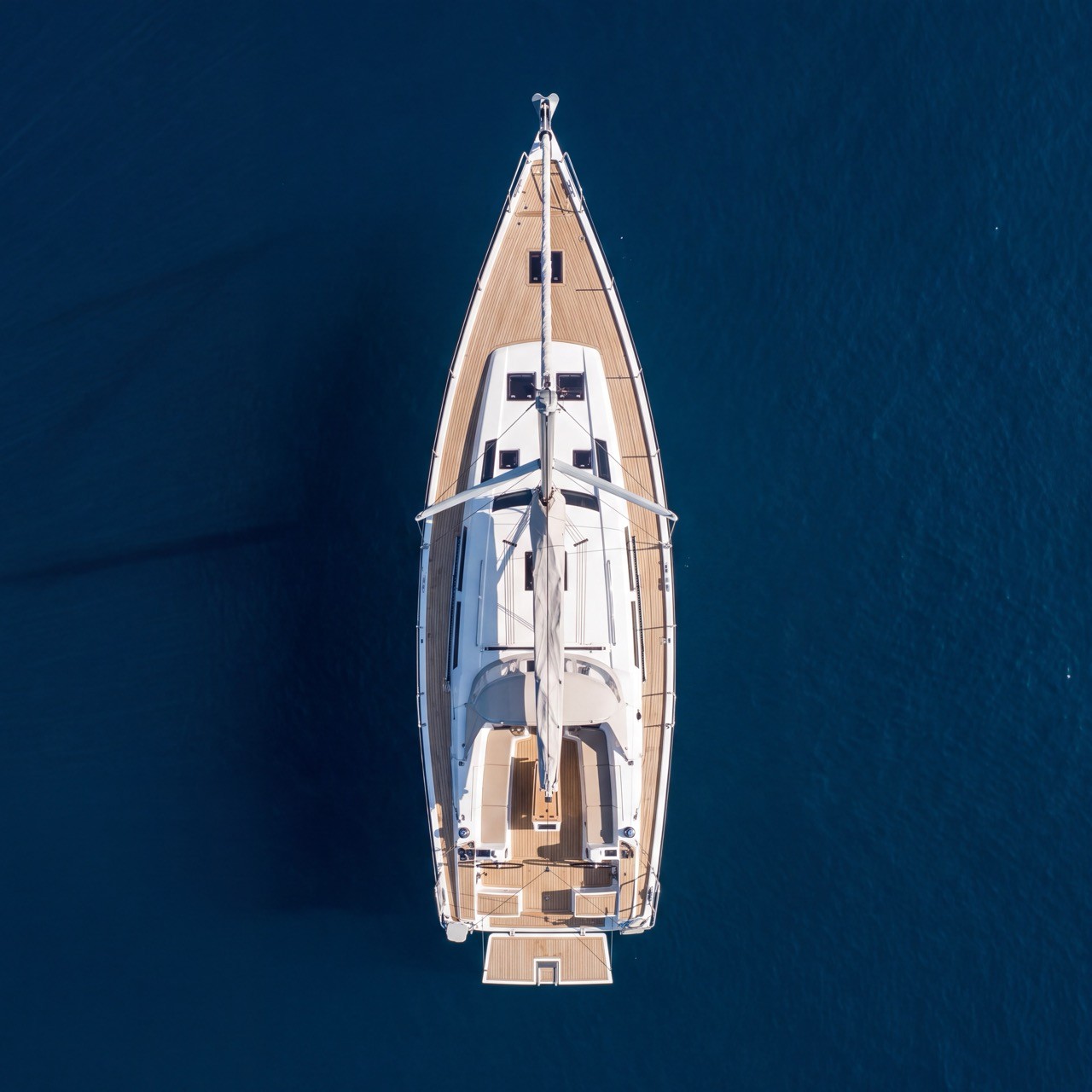 Aerial view looking straight down on a Hanse 575 sailing yacht (2014 model) anchored in deep navy water, showing its long teak-trimmed deck, twin-wheel cockpit, and fold-down swim platform.
