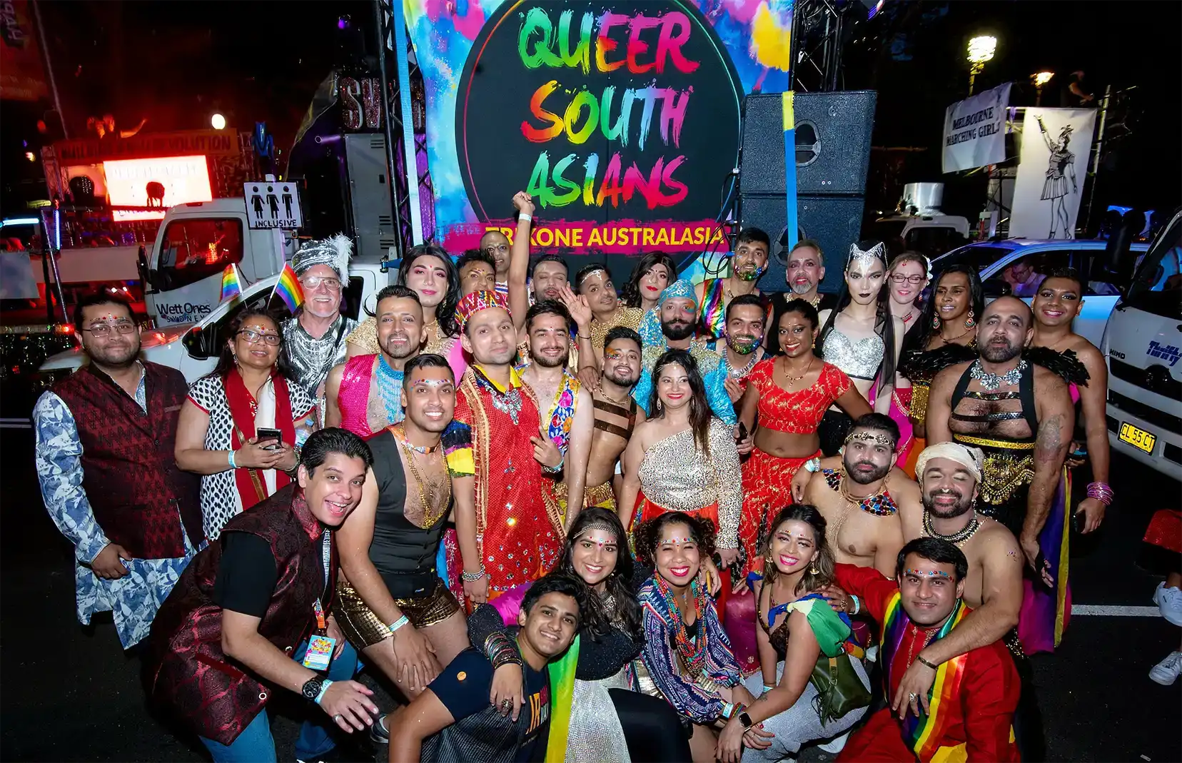 A large, diverse group of people in colourful, festive attire pose together, smiling, in front of a vibrant sign that reads "Queer South Asians, Trikone Australasia" at a lively event or parade.