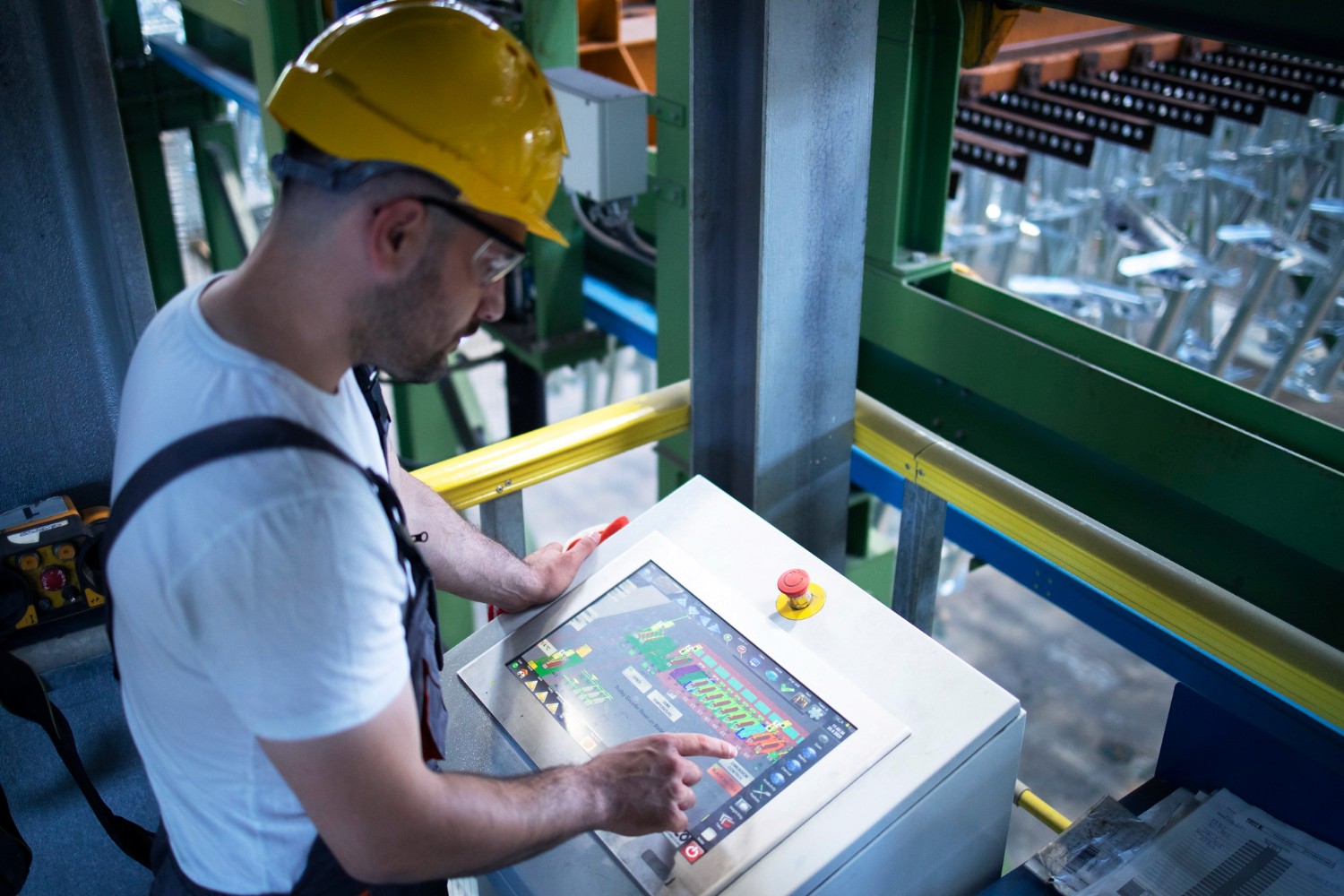 Ein Mann mit gelbem Schutzhelm bedient in einer Industrieanlage ein Steuerpult mit Touchscreen und rotem Not-Aus-Knopf.