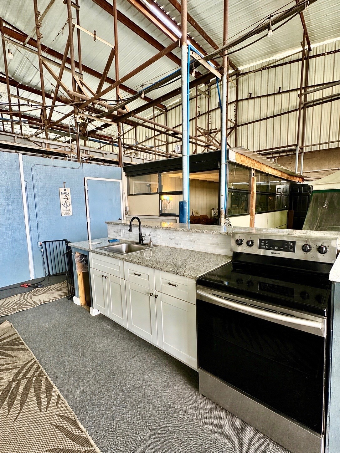 The image shows an industrial-style kitchen featuring a modern black stove, white cabinets, and a sleek granite countertop, set against a background of exposed metal beams and light blue walls.