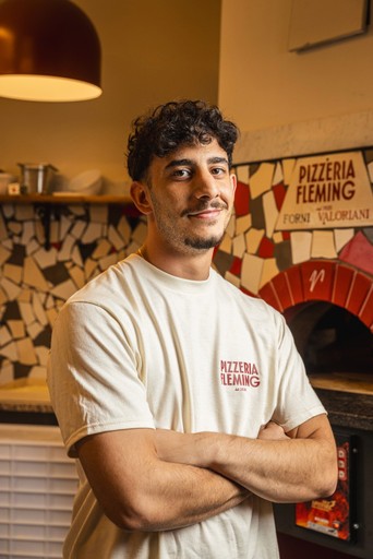A young man stands confidently with arms crossed in a casual setting, with a colorful backdrop of decor.