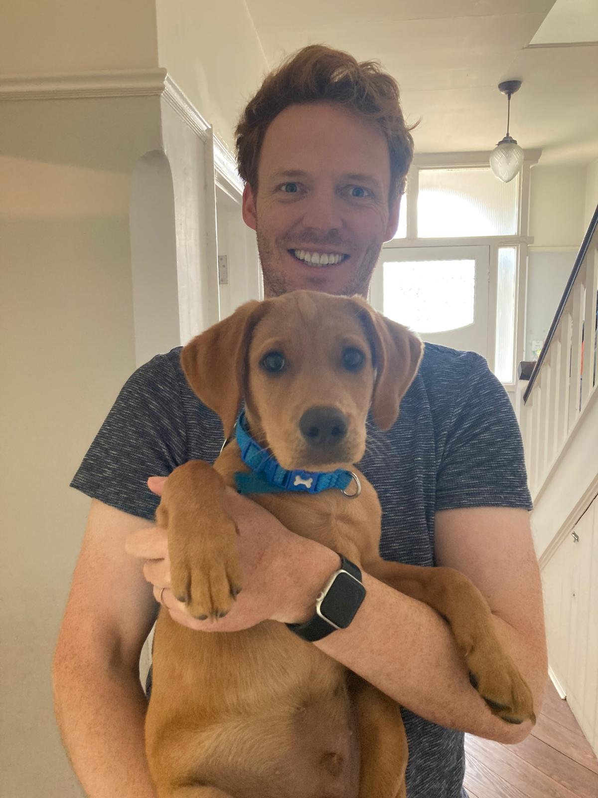 Selfie of a smiling man with red hair holding up a fox red labrador puppy.