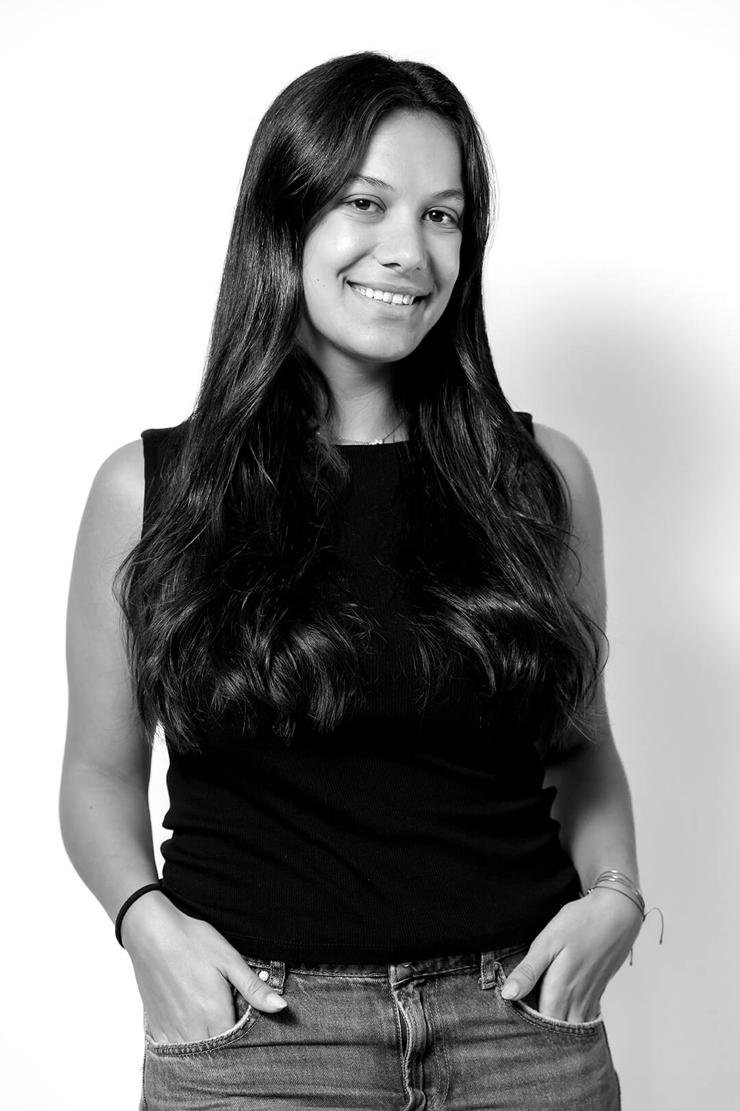 Black and white portrait of a smiling woman with long hair in casual attire, standing with hands in pockets against a plain background. Relaxed and friendly vibe.