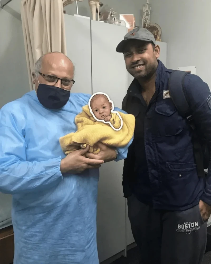Dr. Ashraf Saddik holding a swaddled newborn baby alongside a smiling father at his Melbourne clinic.