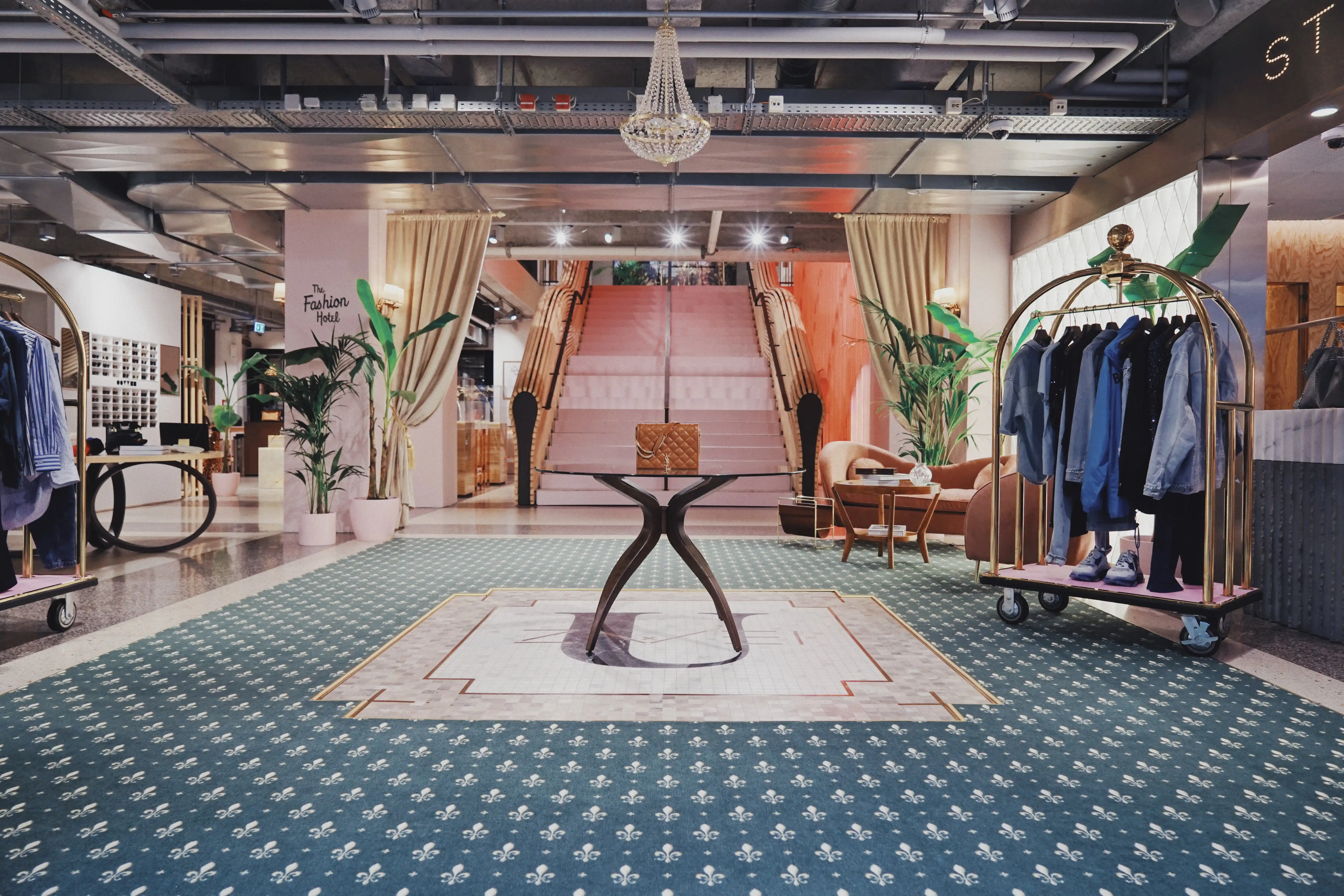 Hotel-themed retail floor at UZWEI FASHION HOTEL with golden luggage trolleys as clothes racks, a glass table with designer handbag, a chandelier above the blush staircase and patterned green carpet.