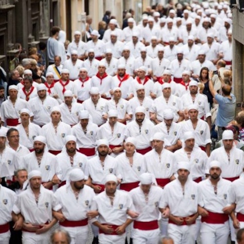 一大群穿著相配白色服裝、系著紅色腰帶和戴著頭巾的人，正沿著狹窄而擁擠的街道行走。