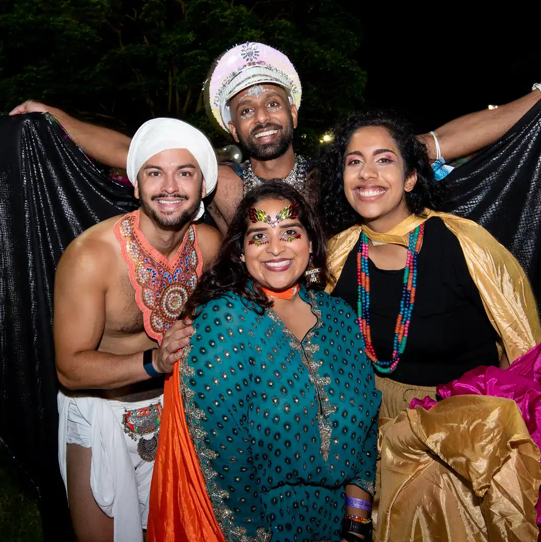 A joyful group of four people in vibrant costumes, beaming at a nighttime outdoor event. Bright colors and cultural attire evoke festivity and celebration.
