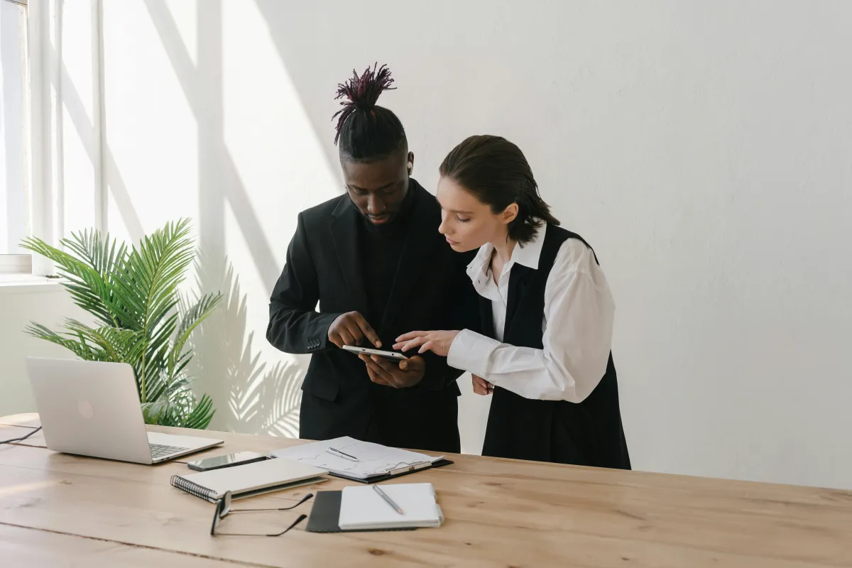 Two professionals, a Black man and a White woman, review a tablet together at a wooden desk, discussing financial strategies.