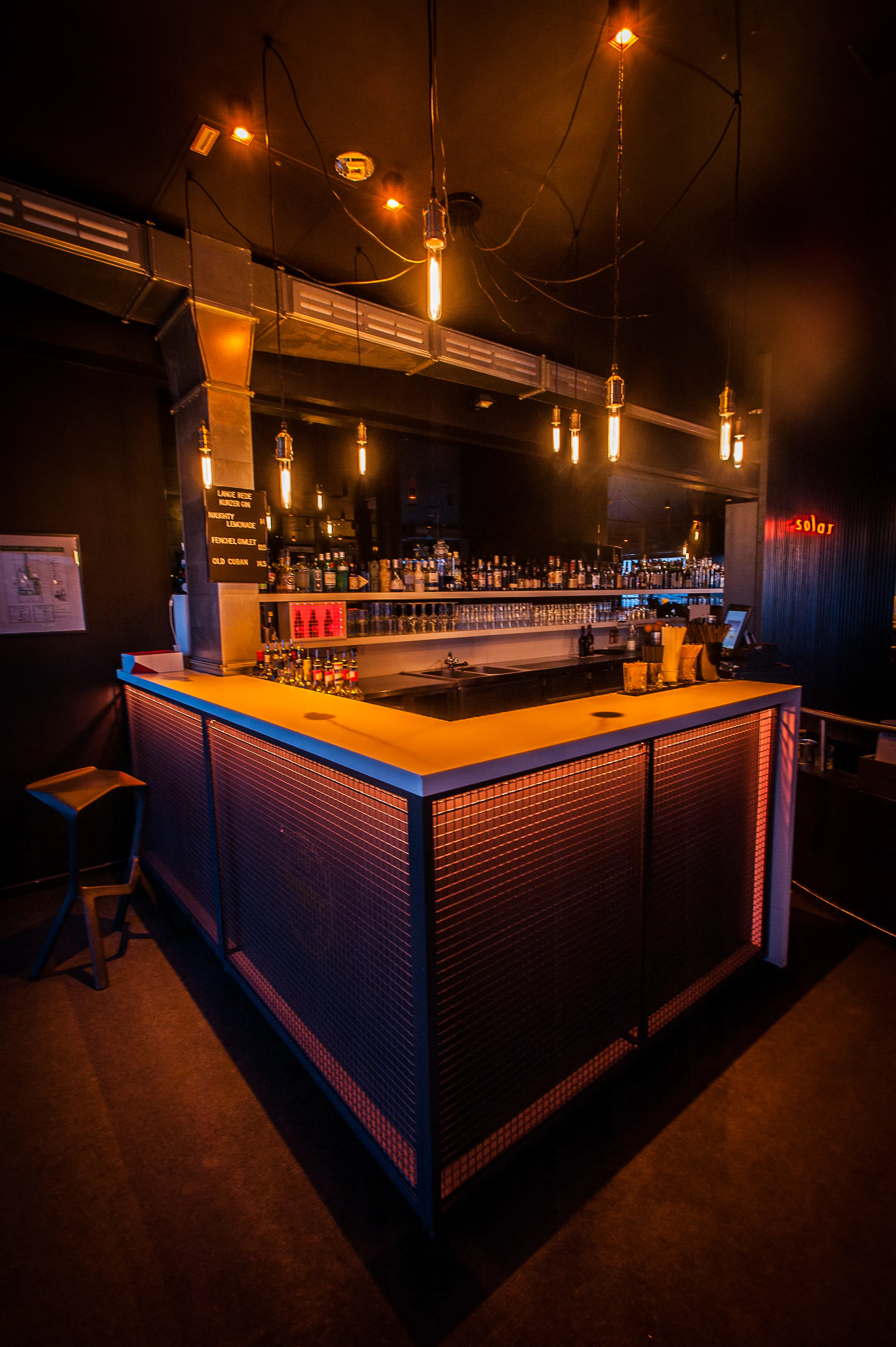 Corner bar at SOLAR SKY LOUNGE with warm-lit mesh front, an illuminated counter top, bottle shelves behind and industrial filament bulbs on black cables.