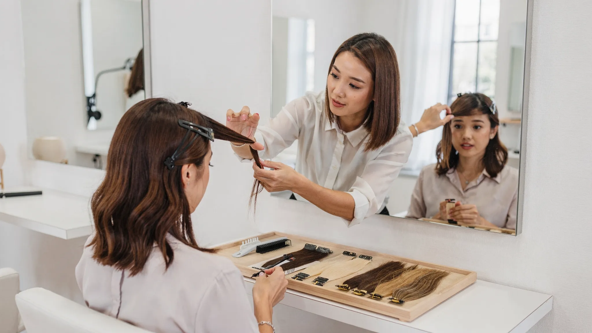 A salon consultation scene with a stylist sectioning a client’s hair while showing a small set of extension samples (tape-in, keratin tip, and weft) on a tray, in a clean, modern salon setting.