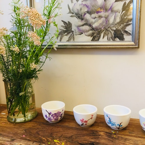 四个花卉图案的杯子放在木桌上,旁边是一个装饰花卉的花瓶,背景是一幅花卉画。