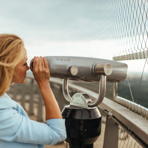 Egy személy érmével működő távcsővel néz egy kilátó teraszról, a háttérben biztonsági kerítéssel és elmosódott tájjal.