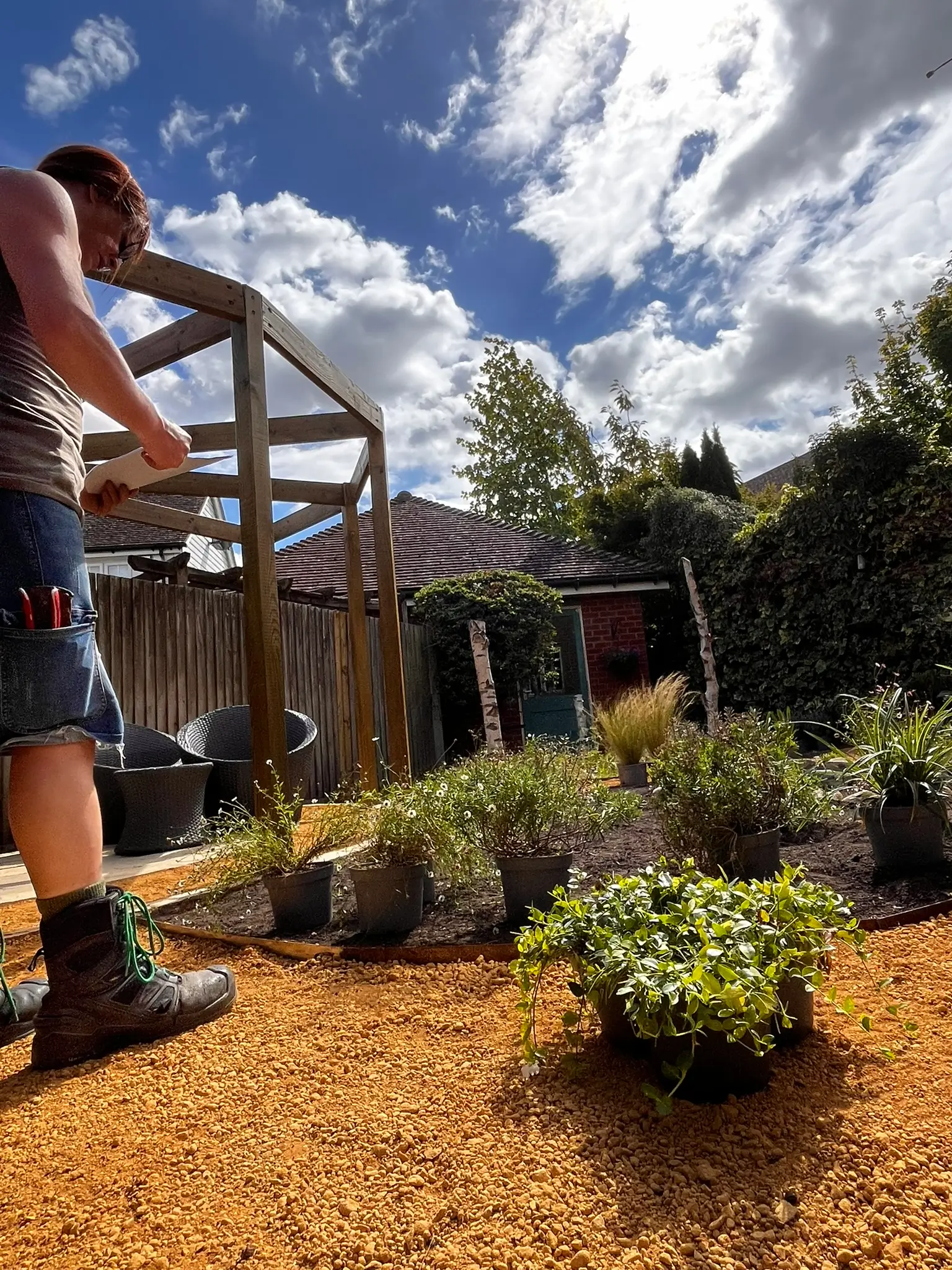 A person tending to plants in a garden under a partly cloudy sky, with wooden structures in the background.