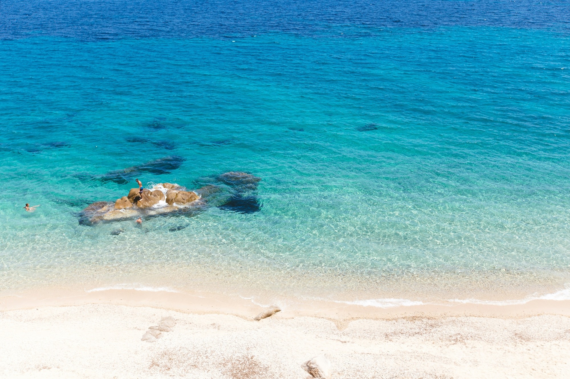 Crystal-clear turquoise sea meeting a white sandy beach with swimmers near rocky outcrops in the Aegean