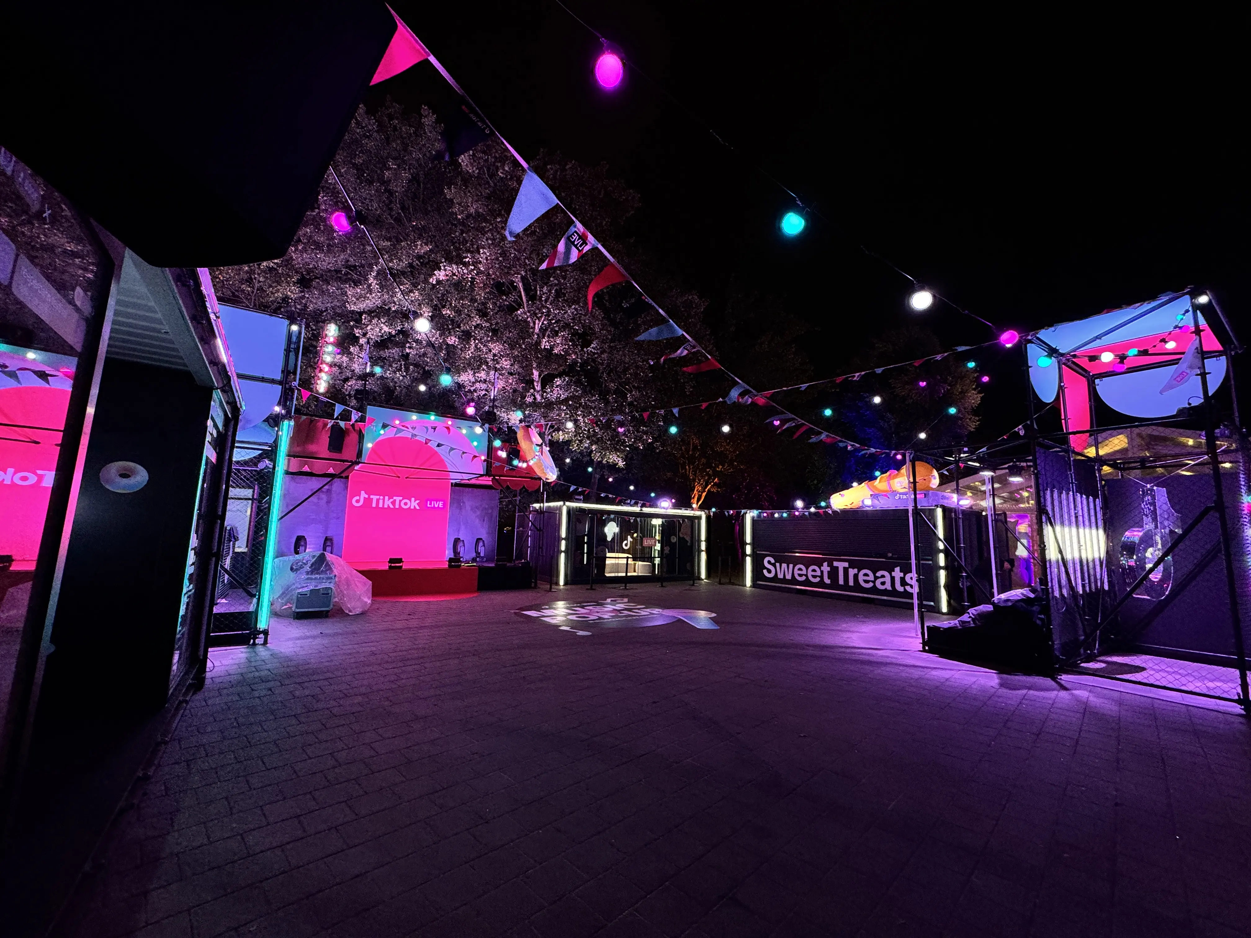 Festival area at night with pennant chains, pink-lit containers and Sweet Treats sign.