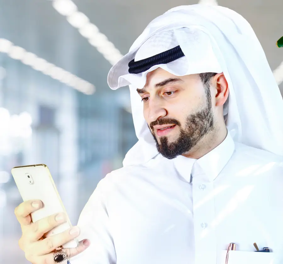 A Middle Eastern man in traditional attire looking at his smartphone, using a digital health app, representing the use of technology in managing chronic conditions like diabetes.