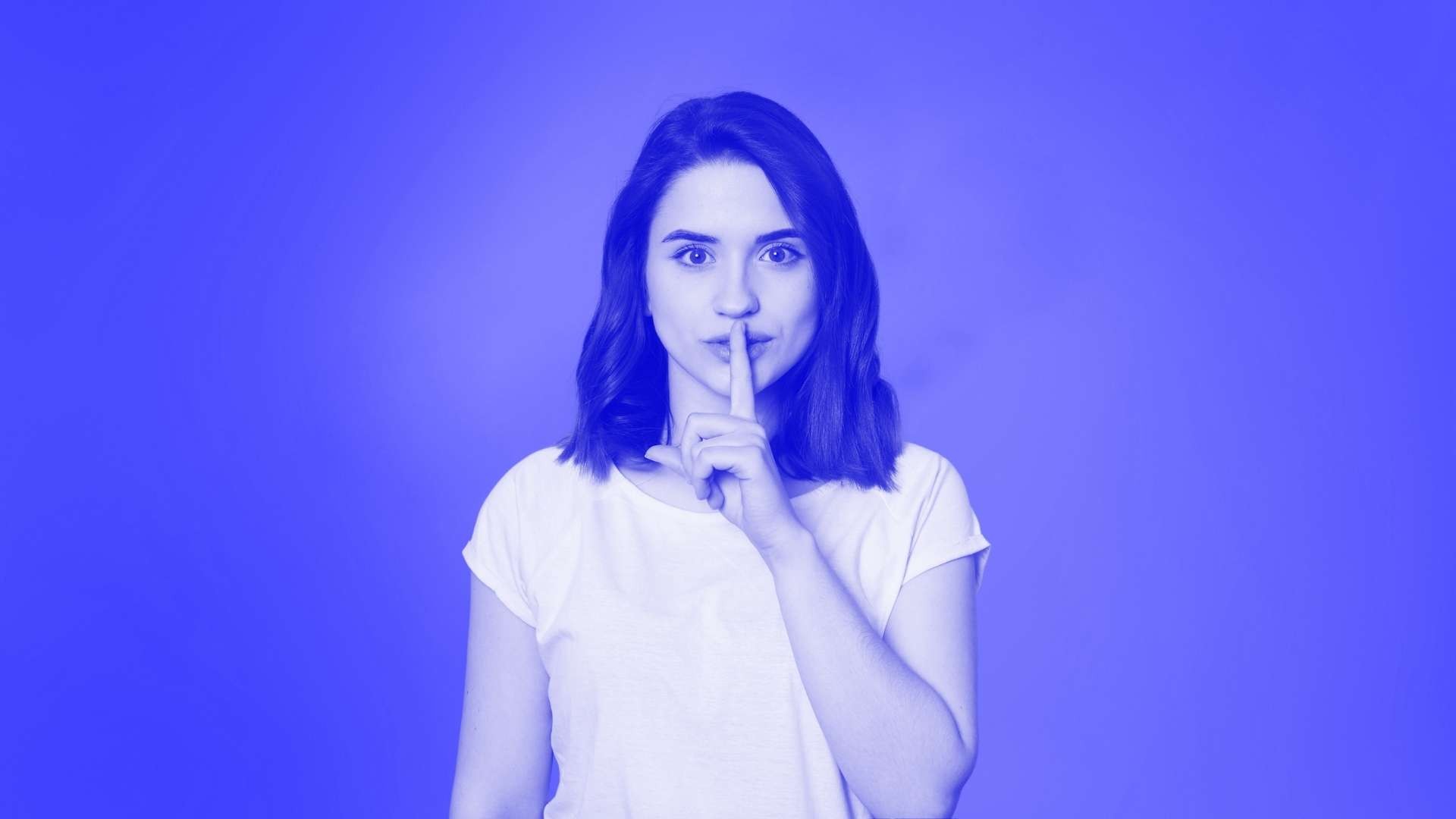 A woman standing looking forward and holding her finger to her lips