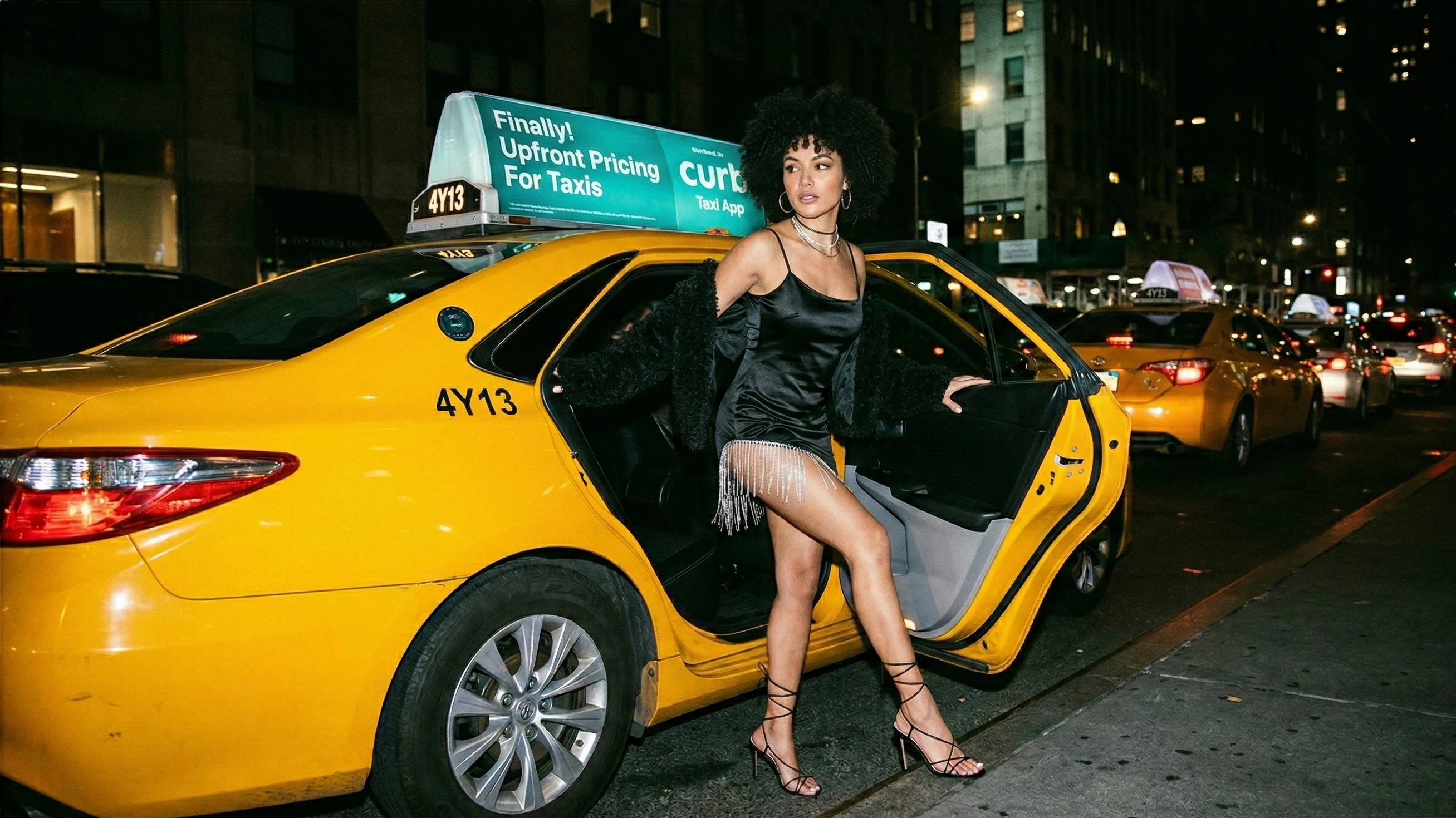 Woman stepping out of a yellow taxi in a black satin mini dress in New York at night.