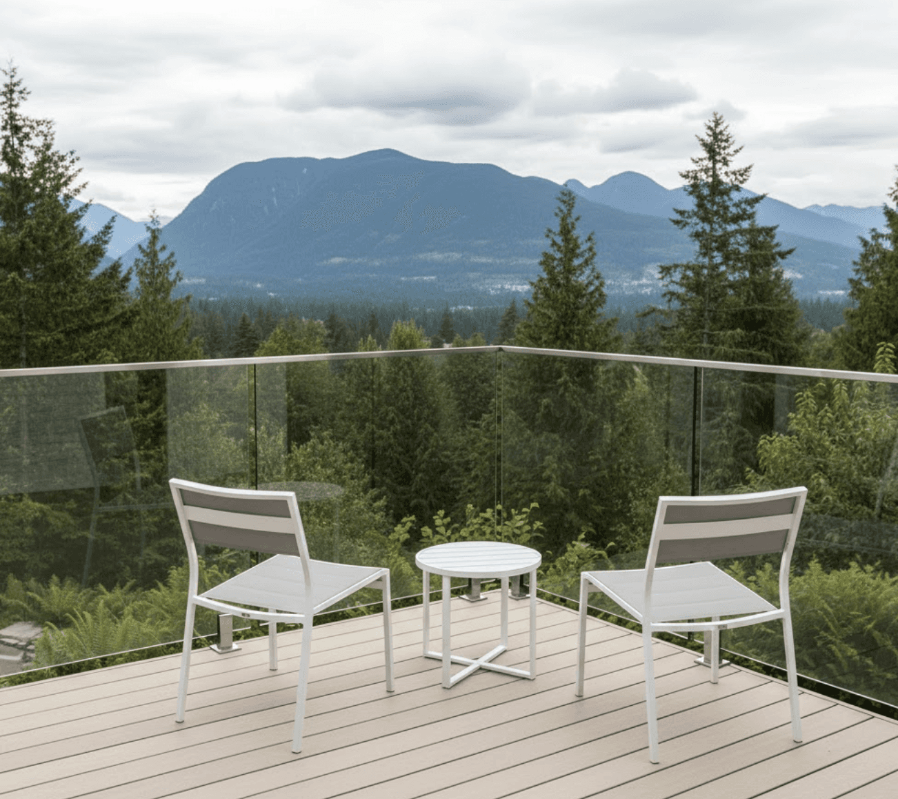 A small modern residential deck approximately 12x12 feet with frameless glass railing system, installed on a Fraser Valley home with stunning mountain views in the background. The deck features light-colored composite decking, clean minimalist design with no visible posts interrupting the glass panels. 
