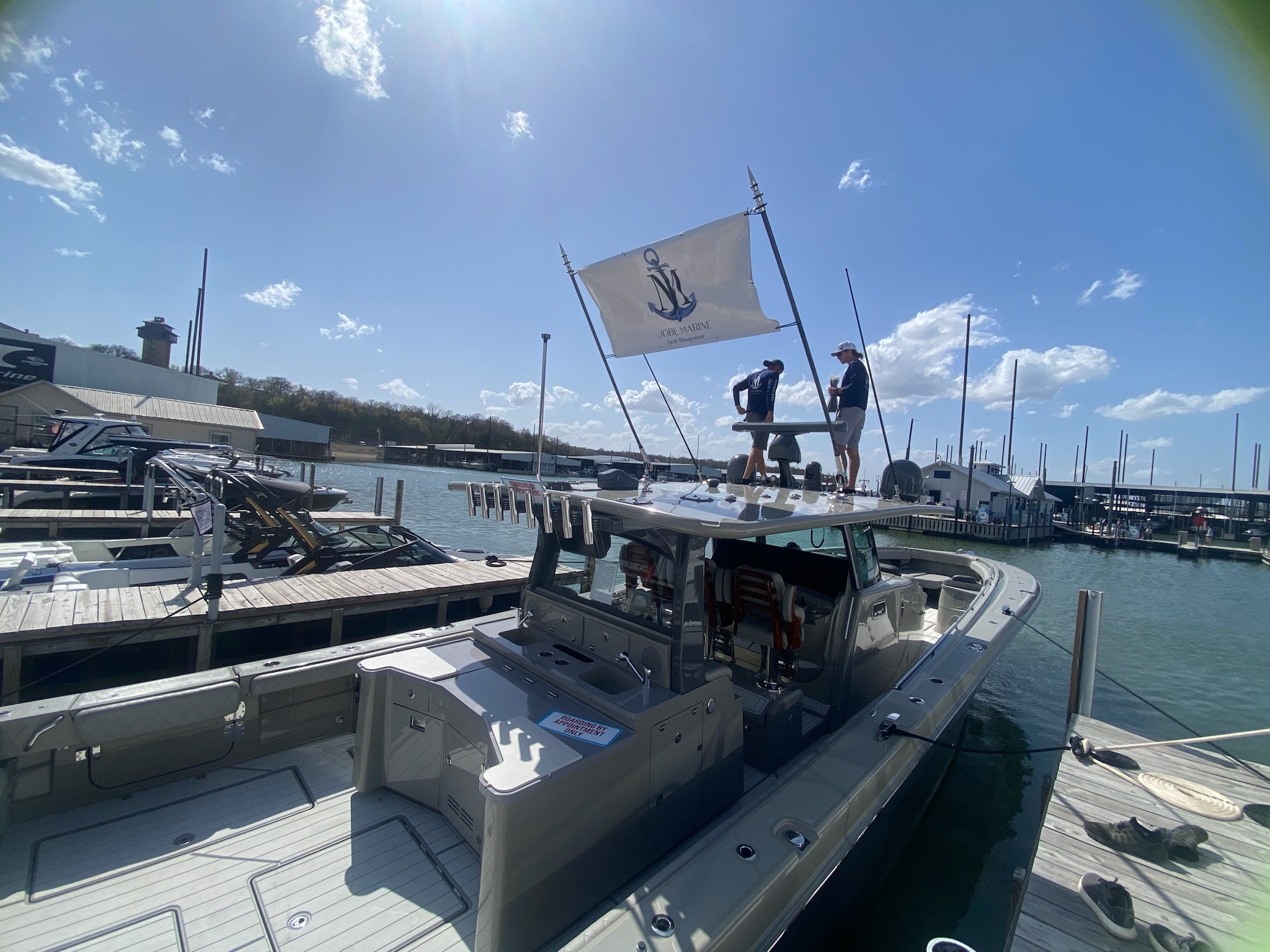 A sleek motorboat docked at a marina under clear blue skies, with two individuals standing on the deck raising a white flag adorned with a blue anchor, surrounded by other boats and a waterfront building.