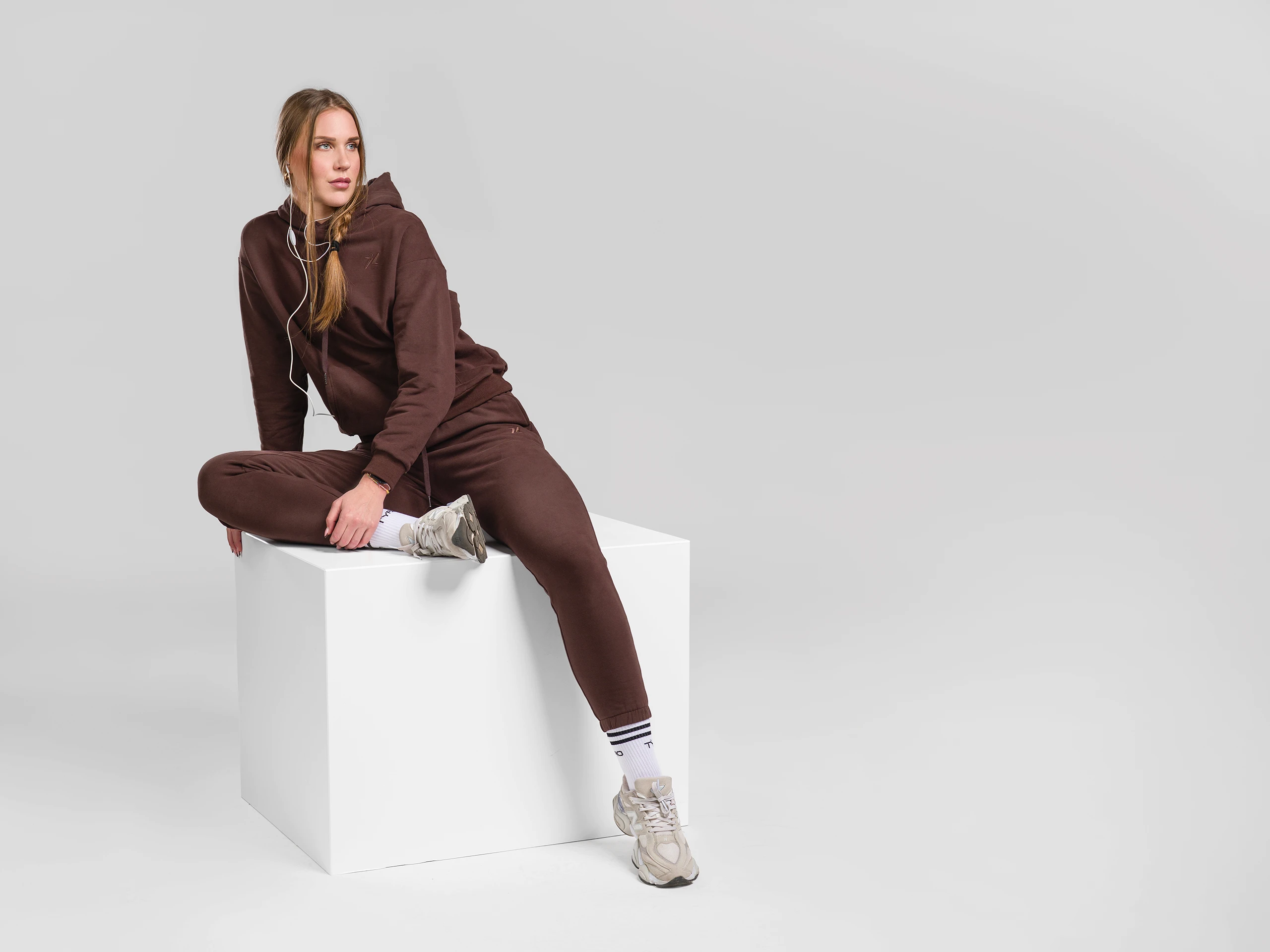 Woman in brown sportswear sitting on a white cube in a studio setting.