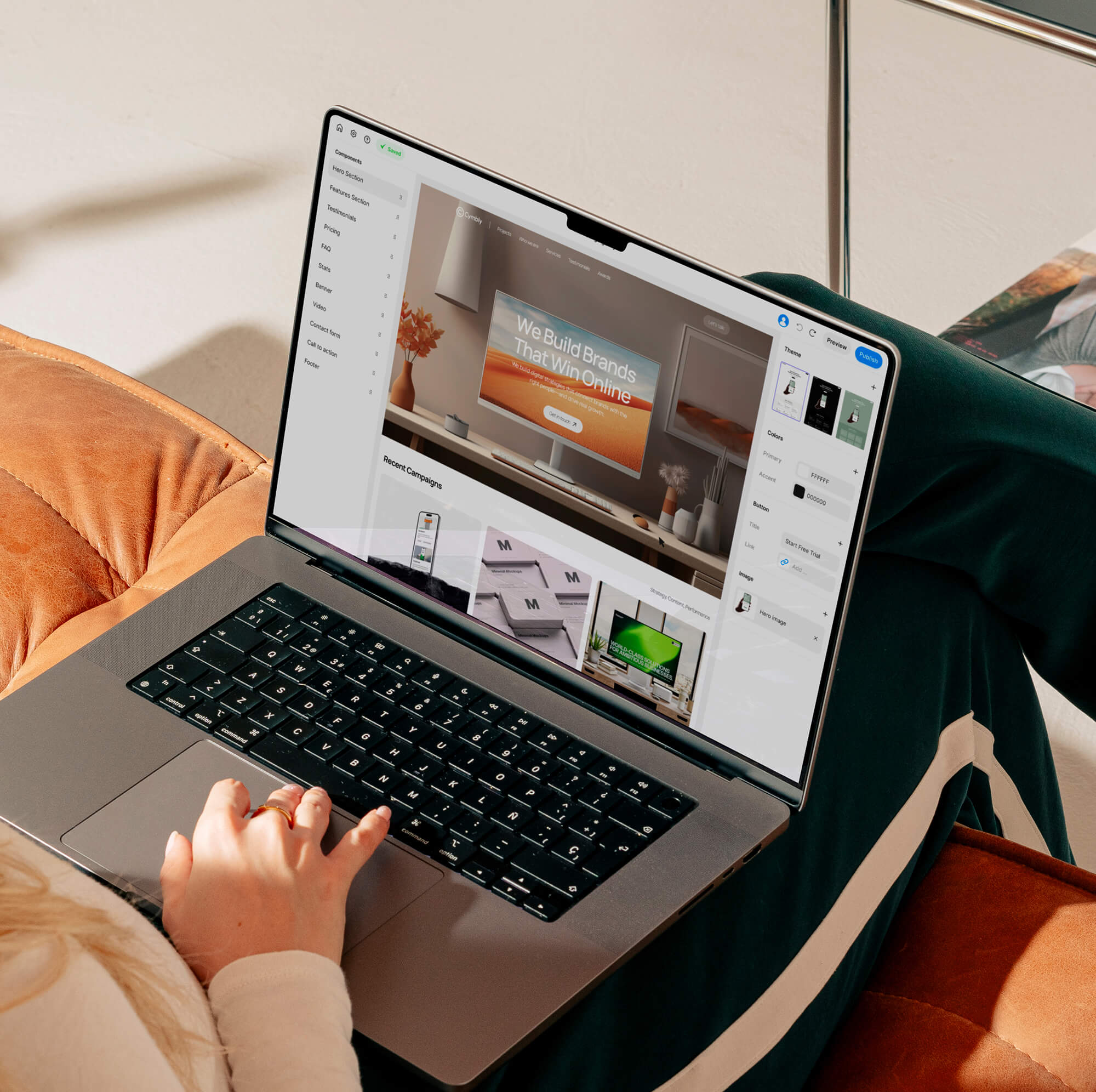 Person using a laptop on a couch, working on a website design in a design application with the screen showing branding and layout elements.