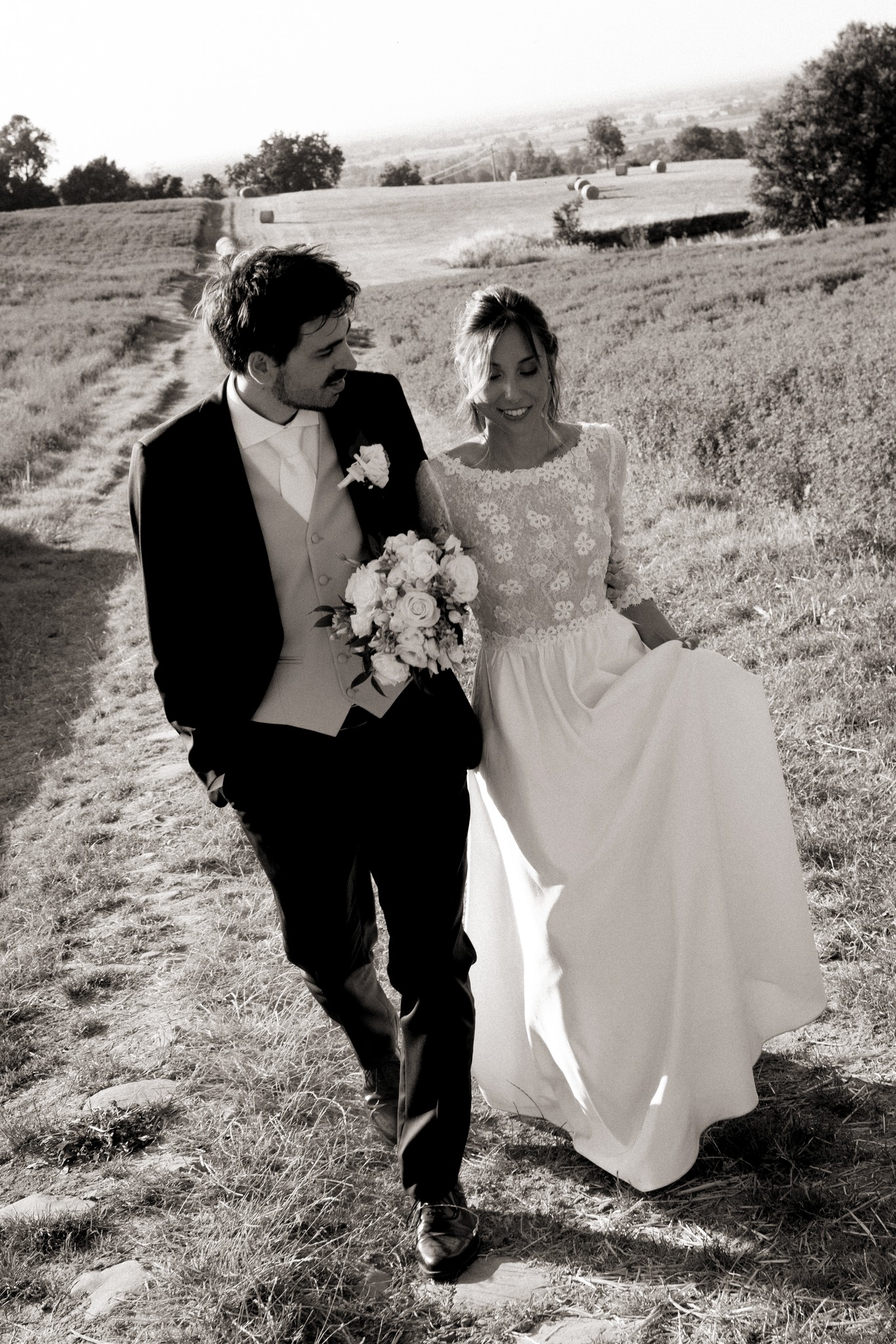 Sposi camminano nei campi di Villa Peroni Riva, lui in smoking nero, lei in abito bianco con bouquet, colline sullo sfondo
