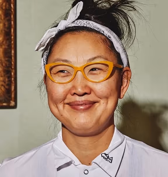 Smiling woman with a white bandana tied on her head, yellow glasses, and a white collared shirt.
