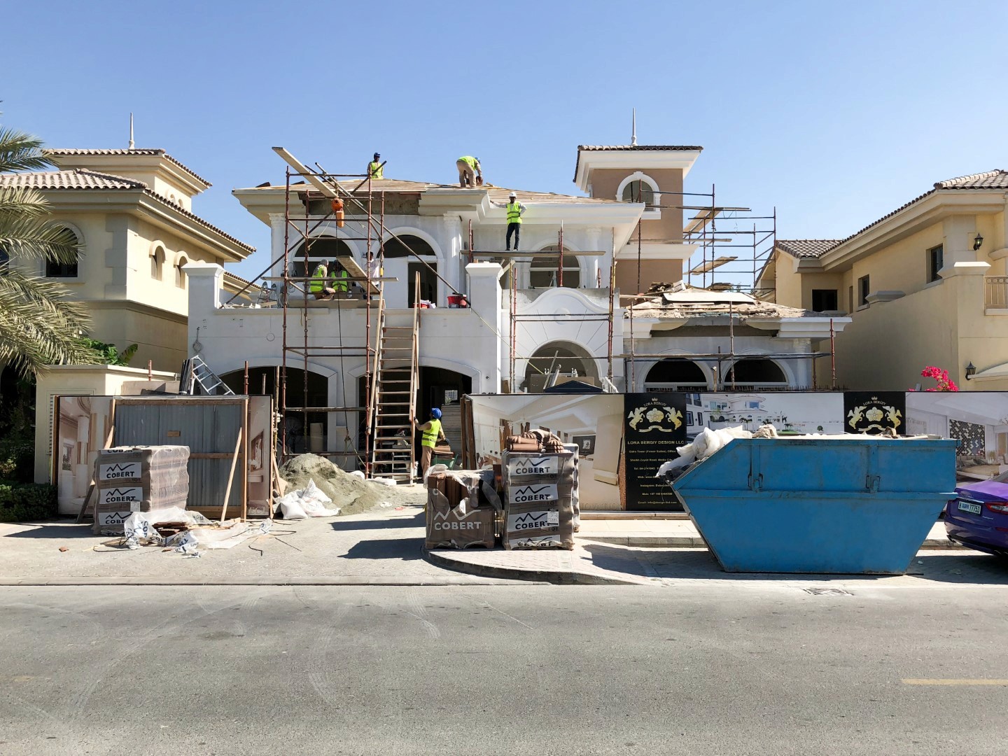 Workers wearing safety gear are renovating a large Mediterranean-style villa with scaffolding, ladders, a skip bin, and construction materials on a sunny day.
