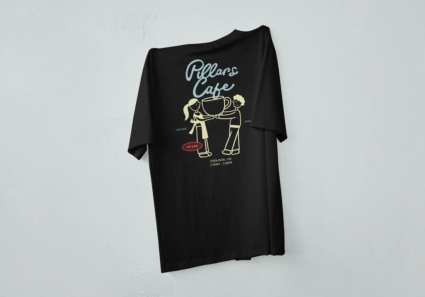 Black Pillars Cafe T-shirt with an illustrated coffee-cup design hanging against a white wall.