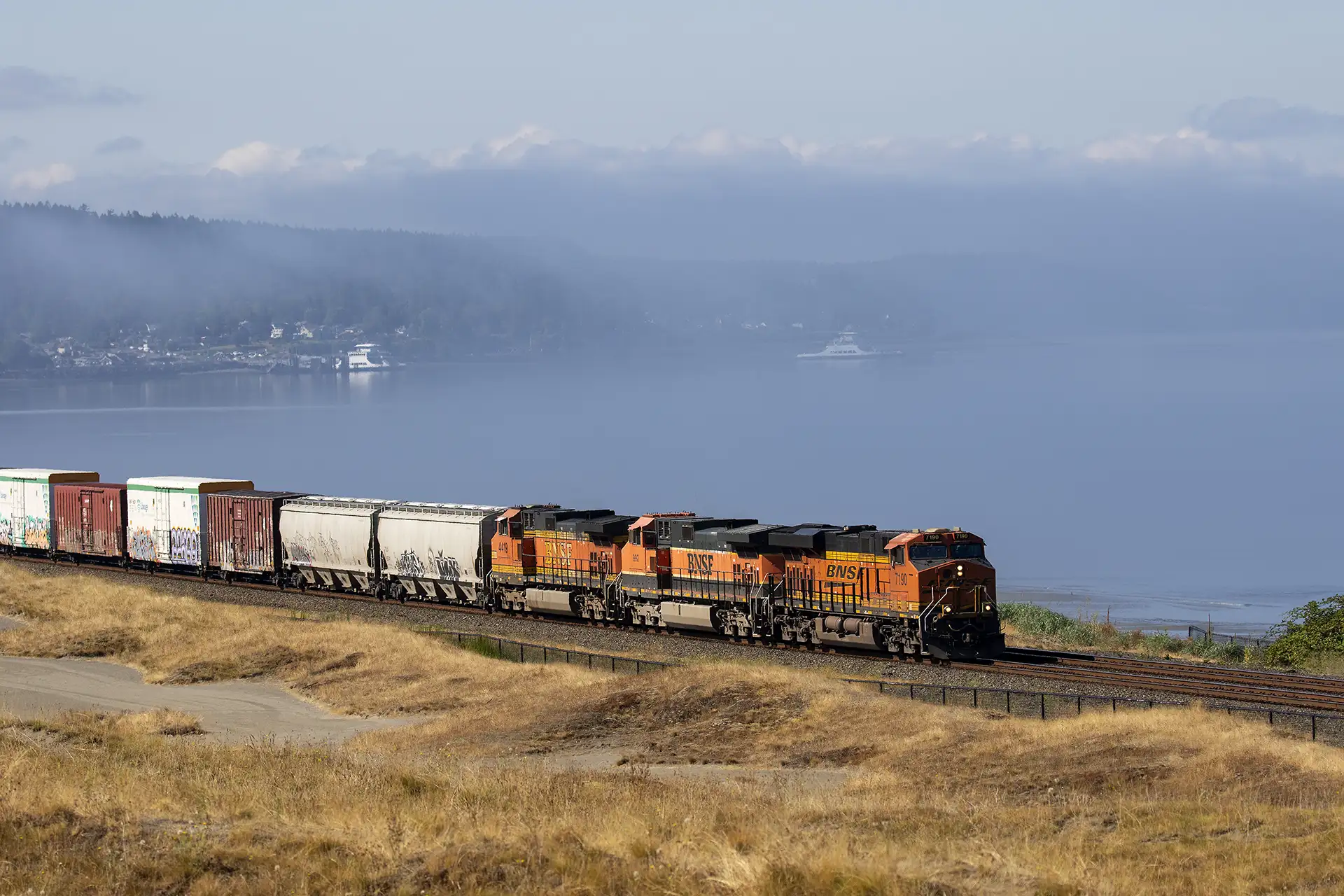 A freight train rolling through hills in the City of University Place. The hills are covered in a yellow grass with spots of bare sand. The Puget Sound sits behind the train, it is a little foggy, and in the distance there are the shores of land with some ships and buildings.
