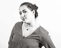 A black and white portrait of a woman with dark hair pulled back from her face. She wears a striped v-neck blouse and a thin necklace with a dark pendant. Her gaze is directed slightly to her left. The background is plain white.