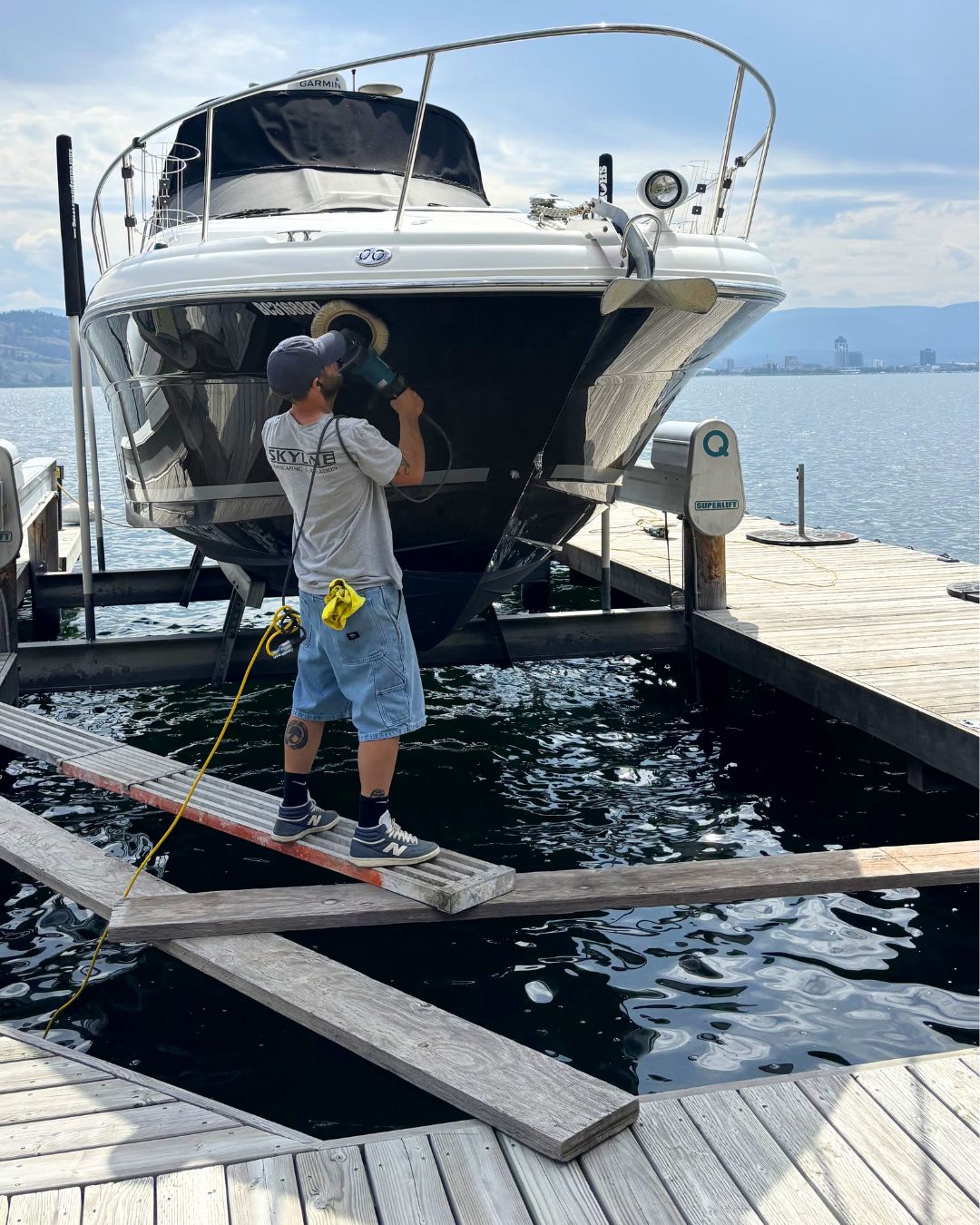 A man working on polishing and detailing the hull of a large boat that is on a dock in a marina out of the water on a boat lift