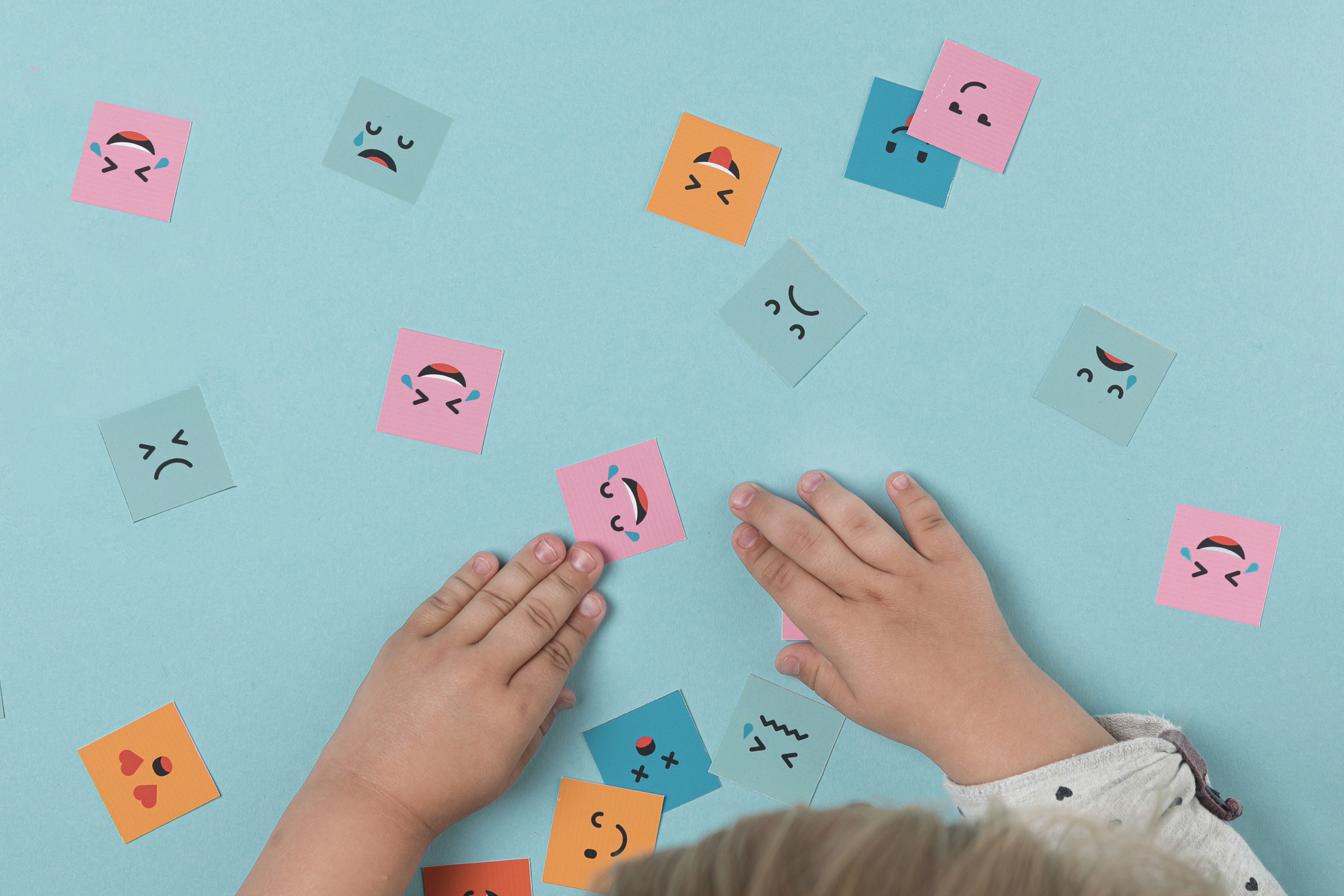 A imagem mostra as mãos de uma criança em uma mesa com várias carinhas de emoções: triste, alegre, piscando etc